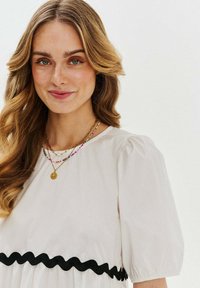 Biele šaty s nafúknutými rukávmi a čiernym vlnitým lemem v páse, vrstvené s viacerými farebnými náhrdelníkmi a zlatým príveskom.