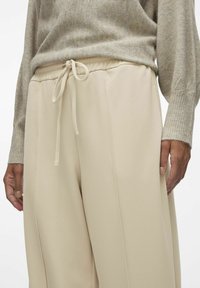 Beige, losse broek met een trekkoord in de taille, gladde textuur en zijnaad. Gecombineerd met een lichtgrijze gebreide trui.