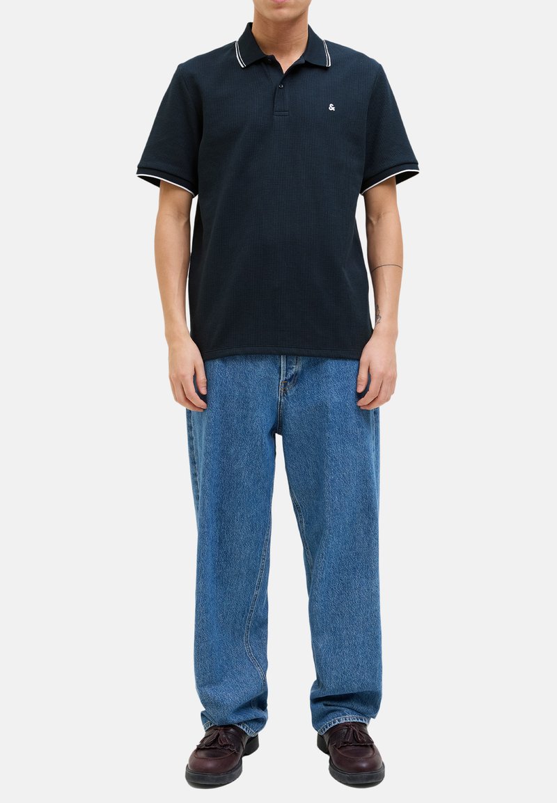 Polo shirt bleu marine avec des bordures blanches, en tissu texturé, avec un col à deux boutons, porté avec un jean bleu ample et des chaussures marron foncé.