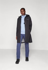 Woman wearing a black hooded coat over a light blue shirt and blue pants, standing with one hand in coat pocket on a plain background.