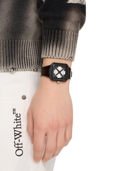 Poignet portant une montre carrée noire avec un motif de croix blanche, associée à un pull en laine gris et un pantalon blanc Off-White.