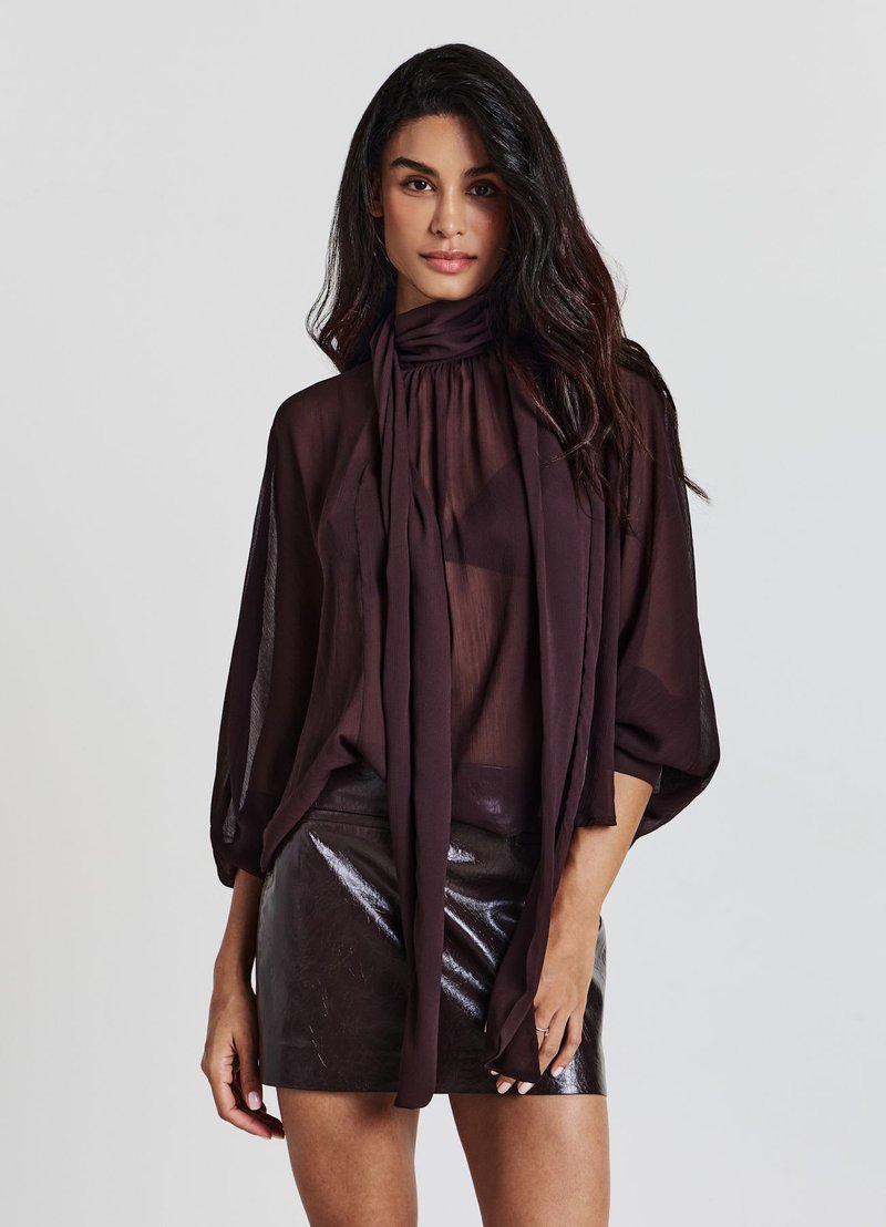 Woman wearing a sheer dark purple blouse with a neck tie and a shiny black leather mini skirt standing against a plain light background.