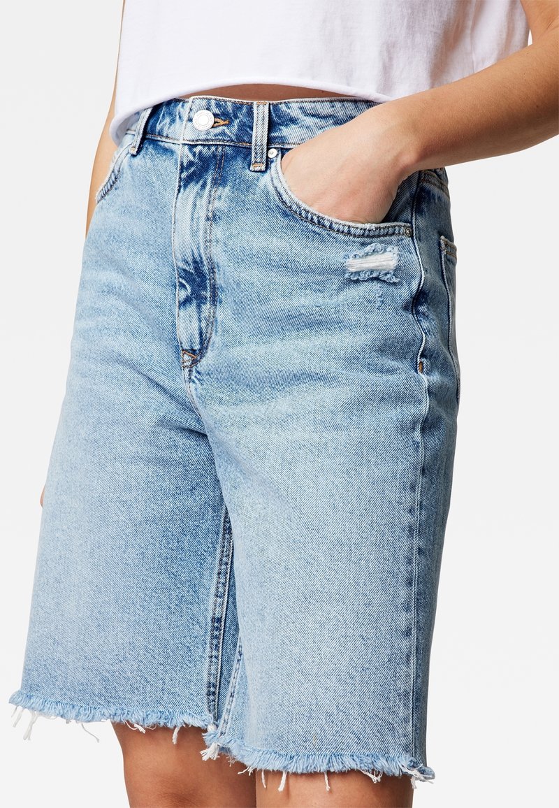 Donna che indossa pantaloni shorts in denim blu chiaro a vita alta con orli sfrangiati e un top corto bianco, con una mano nella tasca anteriore.