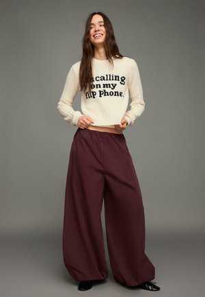 Krämfärgad ribbstickad tröja med svart text "I’m calling on my flip phone." Kombinerad med högmidjade, vida vinröda byxor. Svarta skor.