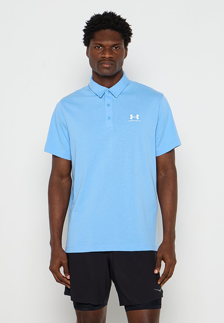Homme portant un polo Under Armour bleu clair et un short de sport noir, debout devant un fond blanc uni.