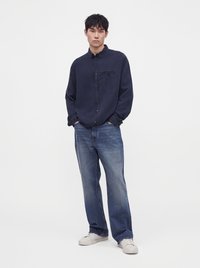 Young man wearing a dark navy button-up shirt, blue jeans, and white sneakers, standing with hands in pockets against a plain background.