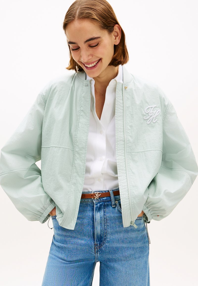 Νεαρή γυναίκα που χαμογελά, φορώντας ένα ανοιχτό πράσινο bomber μπουφάν πάνω από λευκό πουκάμισο, μπλε τζιν και καφέ ζώνη με διακοσμητικά αρχικά.