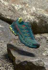 Běžecké boty do terénu, zpevněné tyrkysovou síťovinou, s limetkově zelenými akcenty, červenými detaily a robustní černou podešví s výrazným vzorkem podrážky.