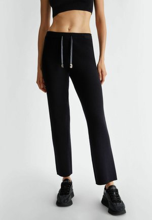 Pantaloni neri in maglia con design a gamba dritta, dotati di vita con lacci e dettagli in metallo argentato. Abbinati a sneakers nere.