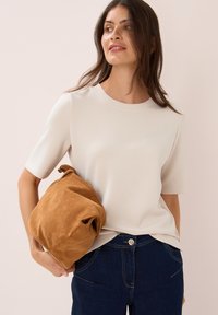 Kvinder med langt, mørkt hår iført en cremfarget kortærmet top og mørke jeans, der holder en stor blød, brun ruskinds- bag under armen.