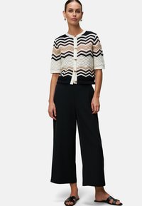Hæklet cardigan i hvid, marineblå og brun zigzag-mønster, korte ærmer, knapfront, parret med sorte bredde-bukser og sandaler.