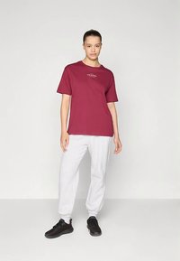 Vīna sarkana kokvilnas T-krekls ar īsām piedurknēm un apaļu apkakli, kombinēts ar gaiši pelēkām sporta biksēm un melniem sporta apaviem.