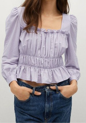 Woman wearing a light purple, long-sleeve, ruched blouse with square neckline and blue denim jeans, hands casually in pockets.