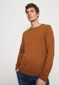 Brun langermet skjorte laget av myk stoff, med rund hals og avslappet passform, brukt med blå jeans, på en nøytral bakgrunn.