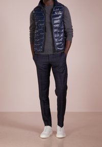 Navy pufová vesta přes šedý svetr, doplněná o tmavé kostkované kalhoty a bílé sneakers. Vesta má zipový přední díl a logo detail.