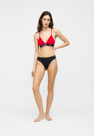 Γυναίκα στέκεται ξυπόλητη φορώντας κόκκινο σουτιέν με μαύρο κείμενο στη λωρίδα και μαύρο εσώρουχο μπικίνι μπροστά σε απλό λευκό φόντο.
