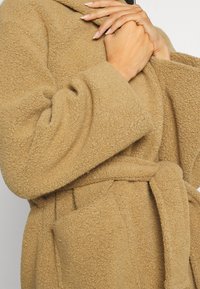 Accappatoio color cammello, morbido, con superficie testurizzata, maniche larghe, vita con cintura e due tasche frontali. Rifinito con un colletto a risvolto.