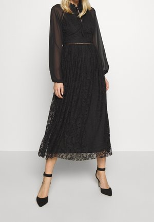 Czarna sukienka midi z koronkową nakładką, długimi przezroczystymi rękawami i dopasowanym gorsetem. Zapięcie pod szyją na kokardę oraz rozkloszowana spódnica. Podkreślone czarne szpilki.