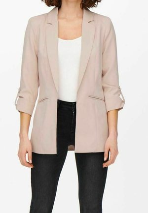 Mujer con una blazer beige claro con las mangas remangadas sobre una camiseta de tirantes blanca acanalada y vaqueros pitillo negros, de pie frente a un fondo blanco.