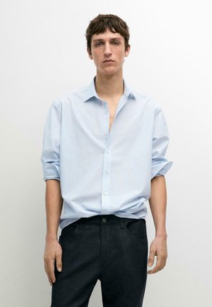 Jeune homme aux cheveux bruns courts portant une chemise rayée bleu clair à manches retroussées et un pantalon foncé, debout devant un fond blanc.