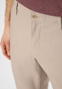 Beige pants with a textured fabric, featuring a front pocket, a visible button closure, and subtle stitching details.