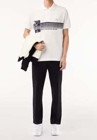 Camisa polo blanca con diseño gráfico azul y rayas, combinada con pantalones negros y zapatillas blancas, sostenida con una chaqueta en blanco y negro.