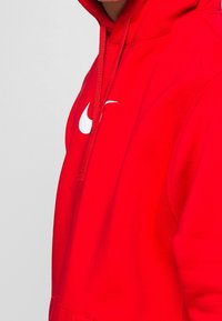 Röd huvtröja i en bomullsblandning med dragsnören, med en stor vit Nike-logotyp på bröstet. Slät textur, avslappnad passform.