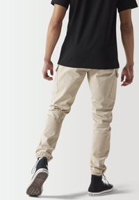 Pantalones cargo beige con puños elásticos y bolsillos laterales, combinados con zapatillas negras de caña alta y una camiseta negra.
