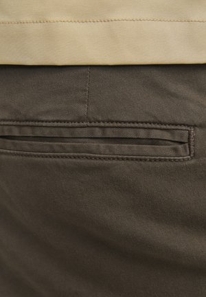 Braune Baumwollhosen mit sichtbarem Gesäßtaschenfeature, akzentuiert durch subtile Nähte und einer glatten Textur. Der Bund ist in einem kontrastierenden Beige.