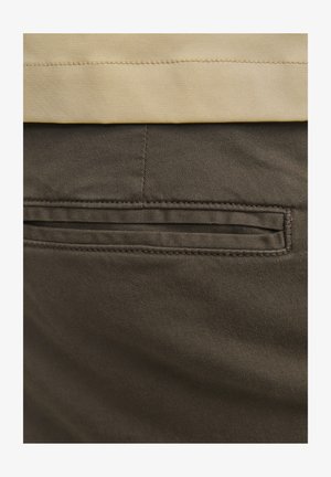 Pruunist puuvillast valmistatud püksid, millel on nähtav tagatasku, kaunistatud peente õmblustega ja siledat tekstuuri. Vöökoht on kontrastse beeži värviga.