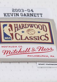Weißes Stoffetikett mit dem Logo "HARDWOOD CLASSICS" in Rot und Blau, mit dem Text "NOSTALGIA CO. Mitchell & Ness" in roter Schrift, Jahr 2003-04 vermerkt.