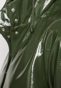Grön blanka regnjacka med en glänsande finish, med silverfärgad hårdvara och en dragsnorskrage för justerbar passform. Slät, texturerad tyg.