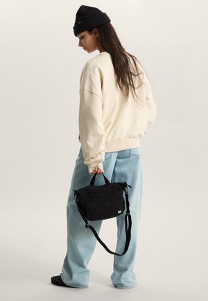 Black fabric bag with dual zippers and adjustable strap, held in hand. Worn with a light-colored sweatshirt and loose denim jeans.