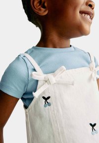 Hvid fløjl overall kjole med blå kirsebærbroderi, parret med en lys blå ribbet t-shirt med korte ærmer. Ribbons binder sig ved skuldrene.