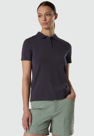 Femme portant un polo à manches courtes sombre et un short vert clair, la main droite dans la poche, sur un fond uni.