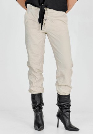 Beige, rynkede bukser med en tilspidset pasform og elastiske manchetter, kombineret med sorte, højhælede, bløde støvler. Sølvknapdetalje synlig.