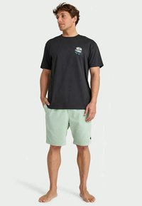 Homem em pé, descalço, a usar uma t-shirt preta com um gráfico de margarida e calções verde-claro, com as mãos nos bolsos, a olhar para o lado.