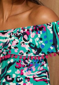 Épaule dénudée portant un haut turquoise à épaules dénudées avec un motif floral abstrait rose, bleu, noir et blanc, et un bord en pompons roses.