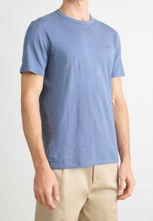 Mand iført en ensfarvet blå t-shirt med korte ærmer og beige bukser, der står mod en hvid baggrund.