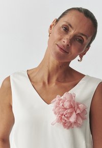 Valkoinen hihaton toppi, jossa on V-kaula-aukko ja vasemmalla puolella tekstuuriempi pinkki kukka-askelma. Samettinen kangas, minimalistiinen tyyli.