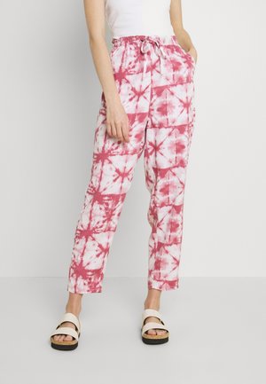 Kvinde iført lyserøde og hvide tie-dye bukser med snøre og lommer, hvid tanktop og hvide platformsandaler, stående mod en hvid væg.