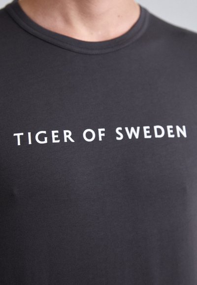 Tamsiai pilkas medvilninis marškinėliai su apvalia kaklo iškirpte, priekyje esančiu baltu tekstu "TIGER OF SWEDEN".