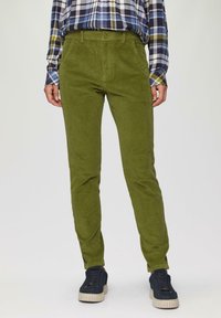 Groene corduroy broek met een slanke pasvorm, met een knoopsluiting en voorzakken, gecombineerd met donkere sneakers. Geruite shirt zichtbaar op de achtergrond.