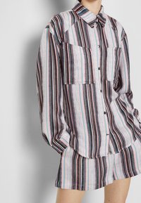Chemise à boutons avec des rayures verticales multicolores en rouge, bleu, noir et blanc. Comprend deux poches sur la poitrine et des manches longues.