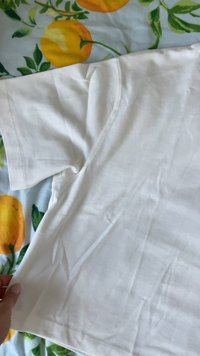 Bela bombažna majica s kratkimi rokavi, z nežno teksturo in klasičnim krojem. Leži na materialu z vzorcem sadnih motivov.