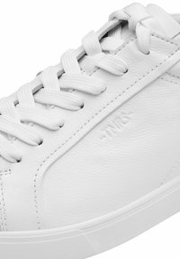 Hvid sneaker lavet af teksturet læder med flade hvide snørebånd, der har et subtilt præget logo og et rent, minimalistisk design.