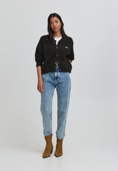 Cardigan negru tricotat cu închidere pe butoane, textură cu coaste și broderie "BALI", purtat peste o cămașă albă și asortat cu blugi albaștri deschisi și cizme maro.