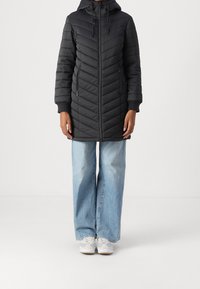 Abrigo largo negro acolchado con capucha, que presenta un cierre frontal, bolsillos laterales y puños acanalados. Combinado con jeans de pierna ancha azul claro y zapatillas.