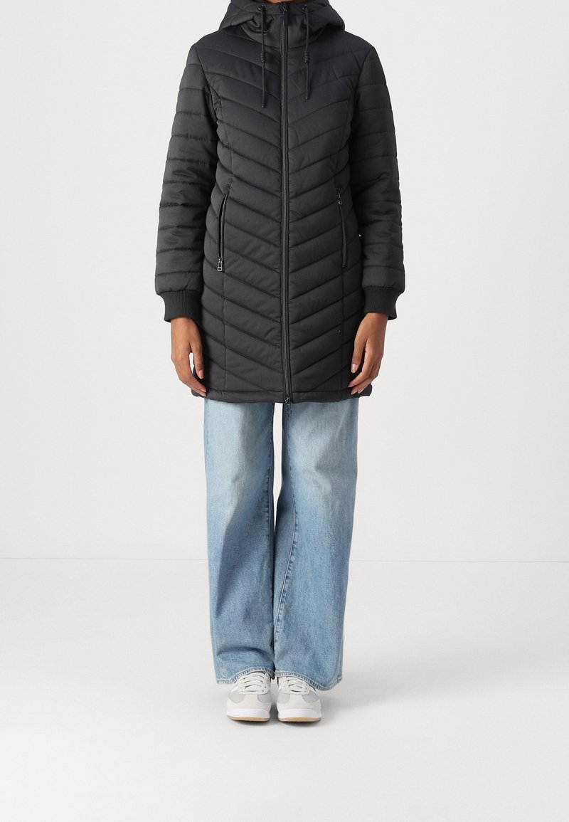 Abrigo largo negro acolchado con capucha, que presenta un cierre frontal, bolsillos laterales y puños acanalados. Combinado con jeans de pierna ancha azul claro y zapatillas.
