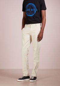 Chinos en coton couleur crème avec une coupe droite, dotés d'une fermeture traditionnelle à boutons et fermeture éclair, assortis à des baskets noires.